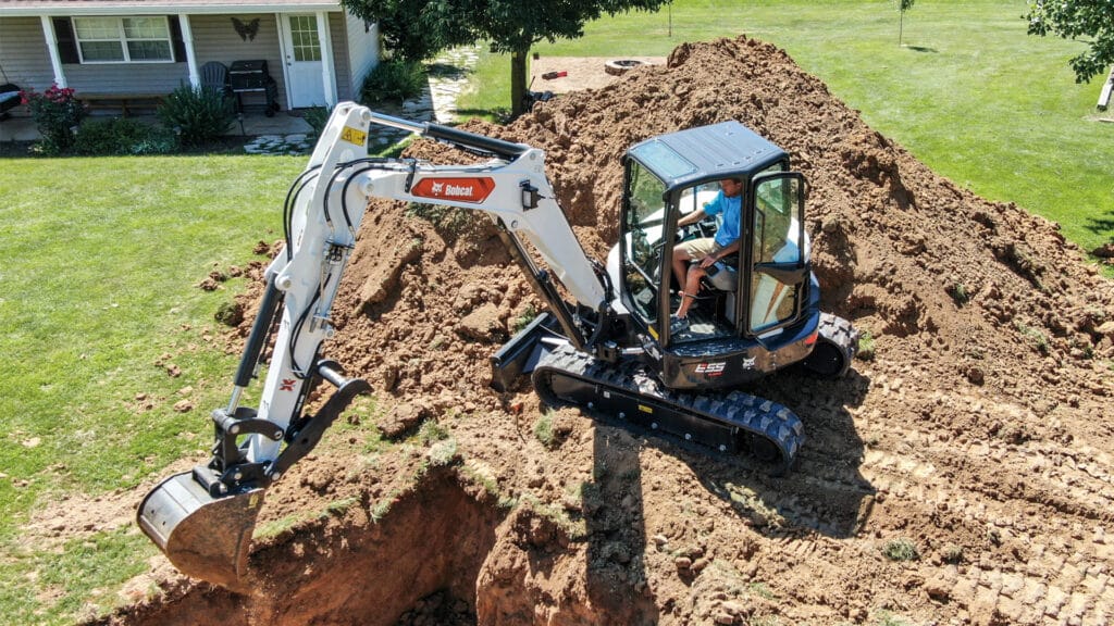 A Bobcat E55R Mini Excavator digging into a residential yard, operated by a worker in a blue shirt, with a large pile of excavated soil and a house in the background. The excavator demonstrates precision and efficiency in a landscaping project.