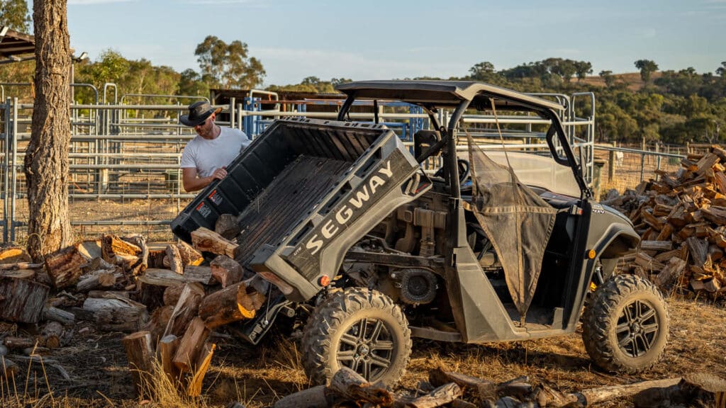 Segway UT6 Navigator UTV unloading a bed full of firewood beside a stockyard, with a man tipping the tray at sunset — ideal for tough rural work and all-terrain hauling.