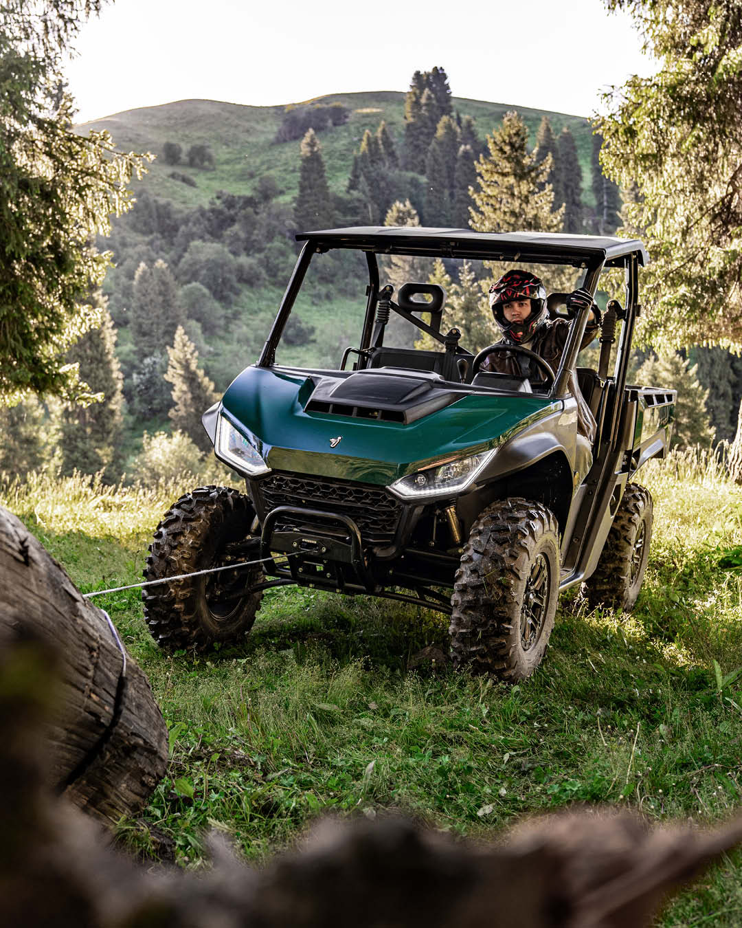 A Segway UT6 Pro UTV using a winch to pull a large log in a forested hillside area, with the driver wearing a helmet and surrounded by lush green trees and grass.