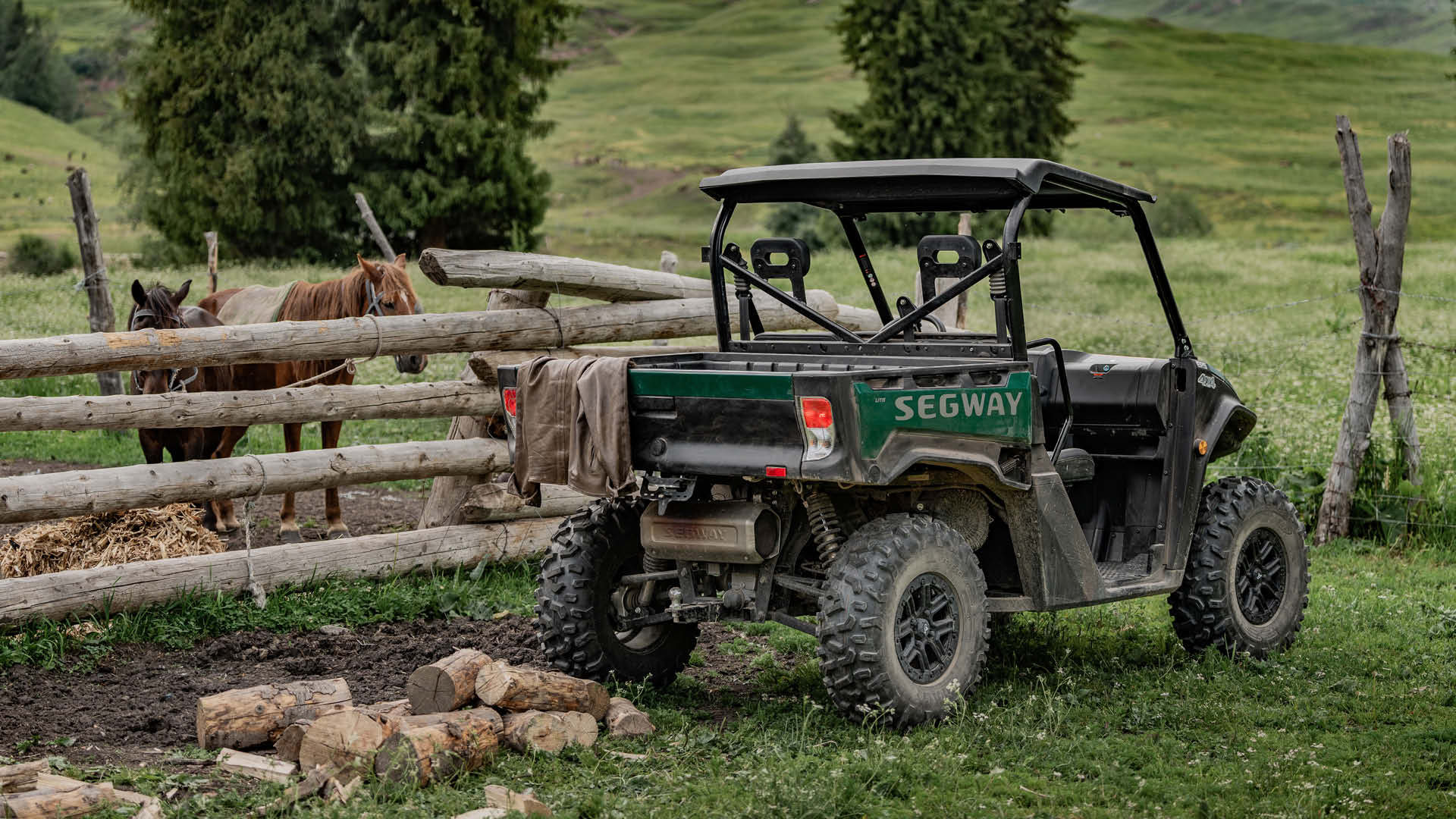 A Segway UT6 Pro UTV parked on grass near a wooden fence with two horses, firewood on the ground, and rolling green hills in the background.