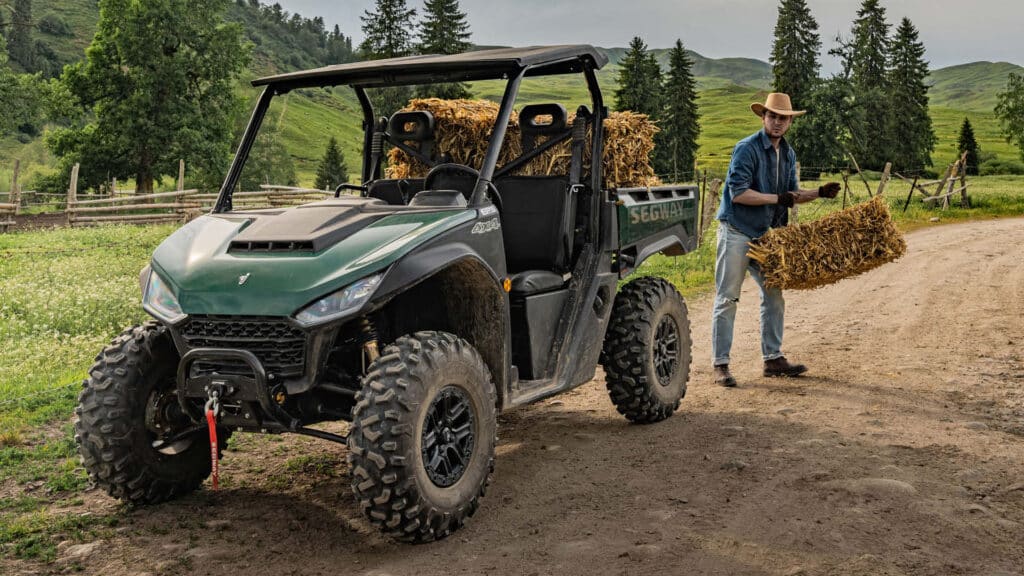 A Segway UT6 Pro UTV loaded with hay parked on a dirt track, while a farmer in a hat unloads a bale in a scenic rural setting with rolling green hills and wooden fences.