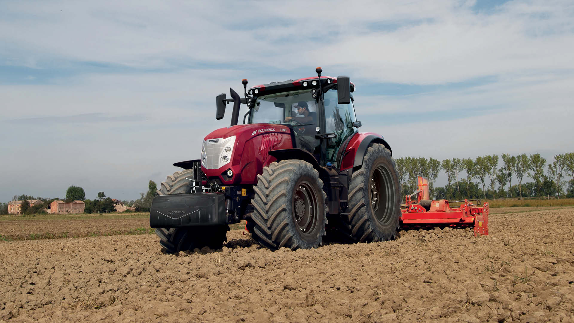McCormick X7 farm tractor working in a dry, ploughed paddock with a red implement attached, driven by a farmer under a clear sky.