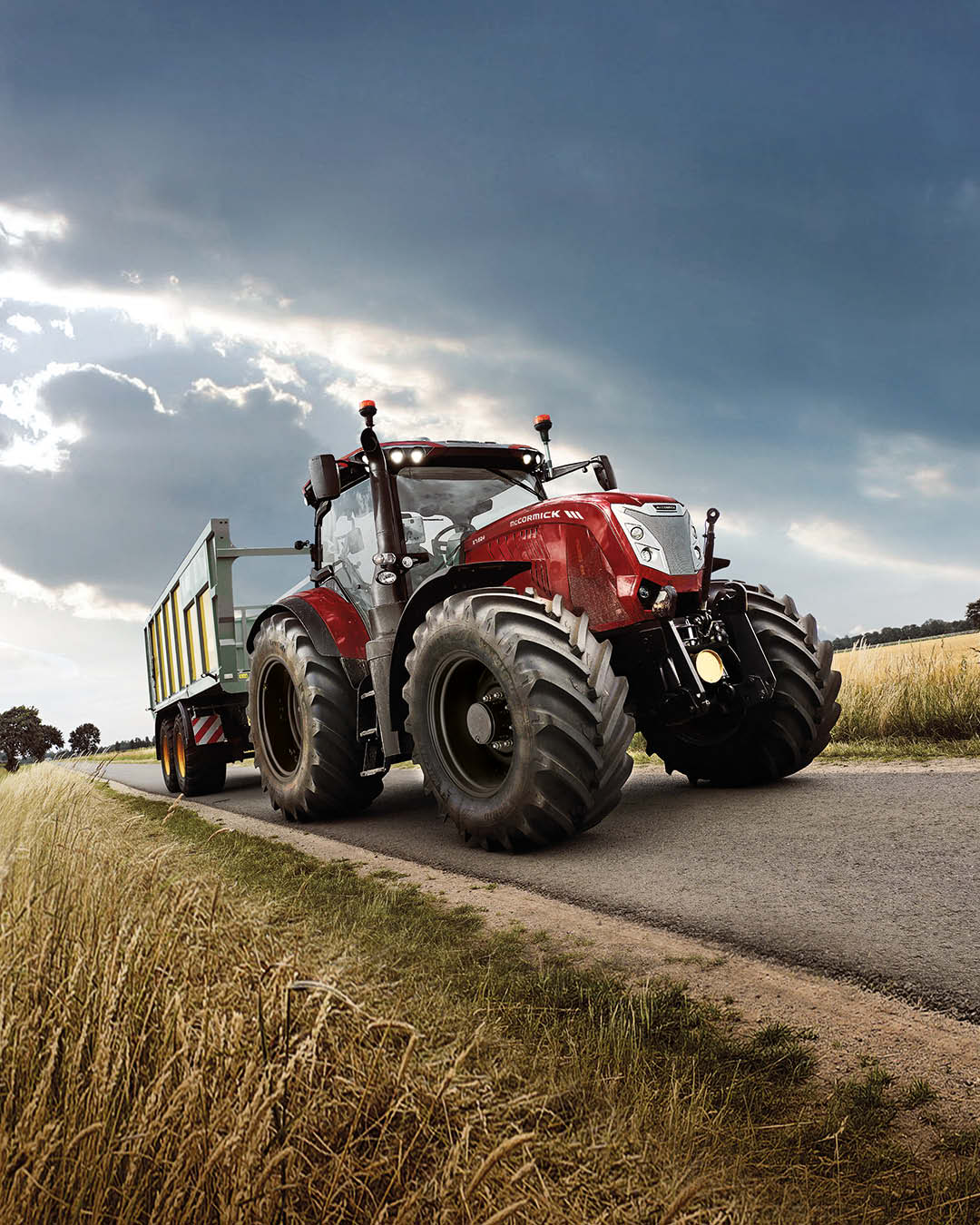 McCormick X7 farm tractor hauling a large green trailer on a country road under a dramatic sky, with wheat fields on either side.