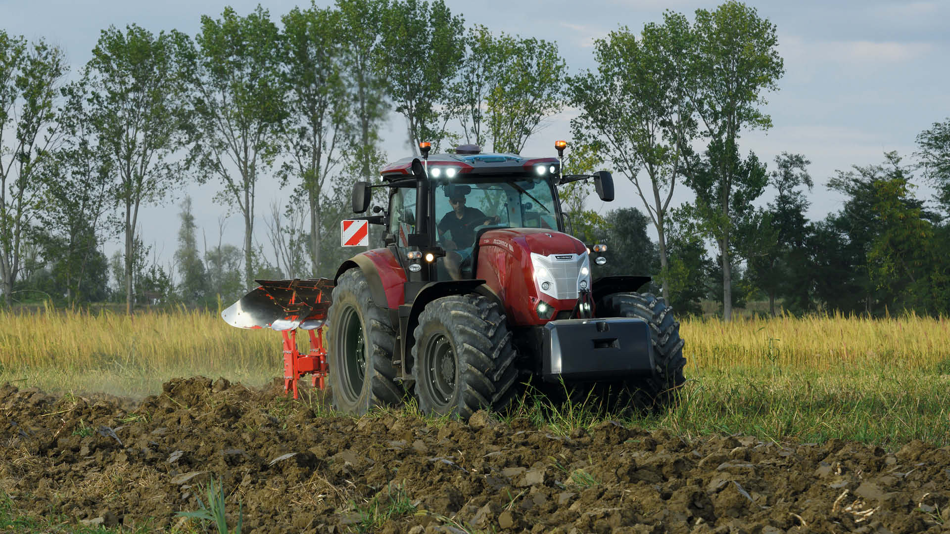 McCormick X7 farm tractor working in a green field, pulling a red plough through freshly turned soil with trees in the background.