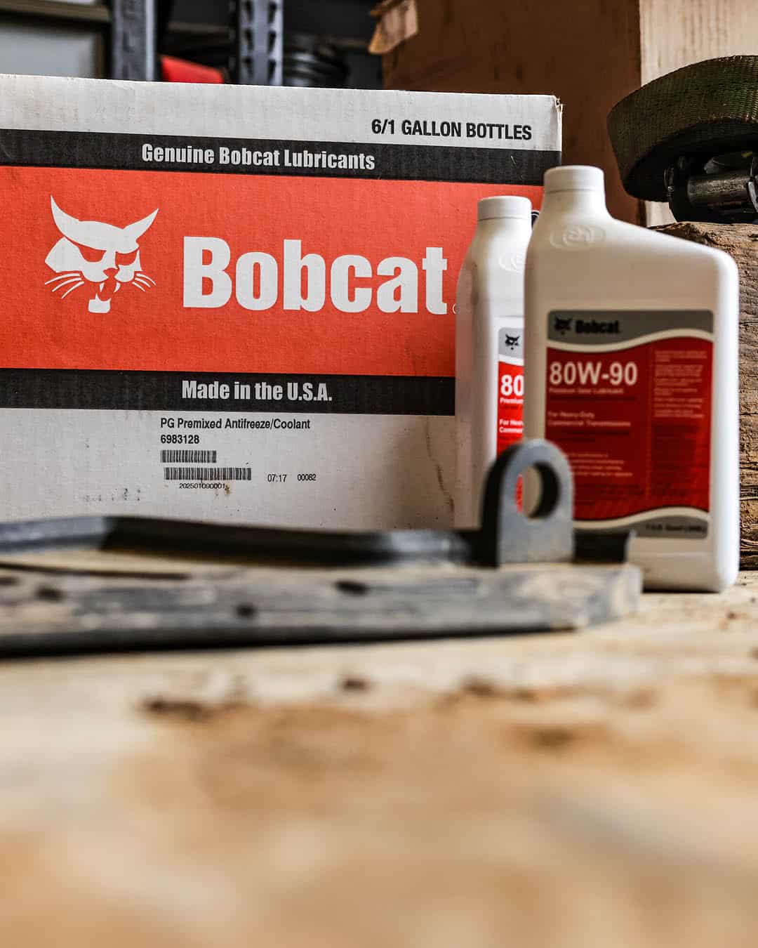 Genuine Bobcat lubricants including 80W-90 gear oil bottles and a boxed shipment of PG premixed coolant displayed in a service workshop setting.