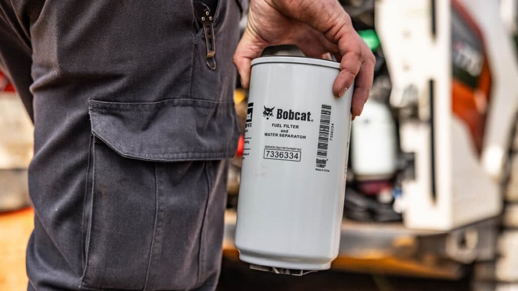A Bobcat fuel filter and water separator held by technician in workshop near equipment.