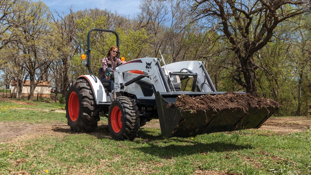 Woman operating a small tractor for sale with front-end loader, moving soil on a hobby farm surrounded by trees.