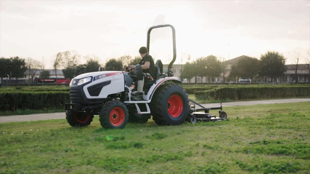 Man using a small tractor for sale with slasher attachment to maintain a grassy field near a rural property.