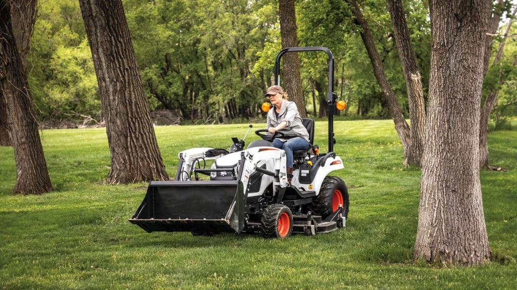 Woman operating a small tractor for sale with front-end loader attachment, navigating through trees on a green hobby farm.