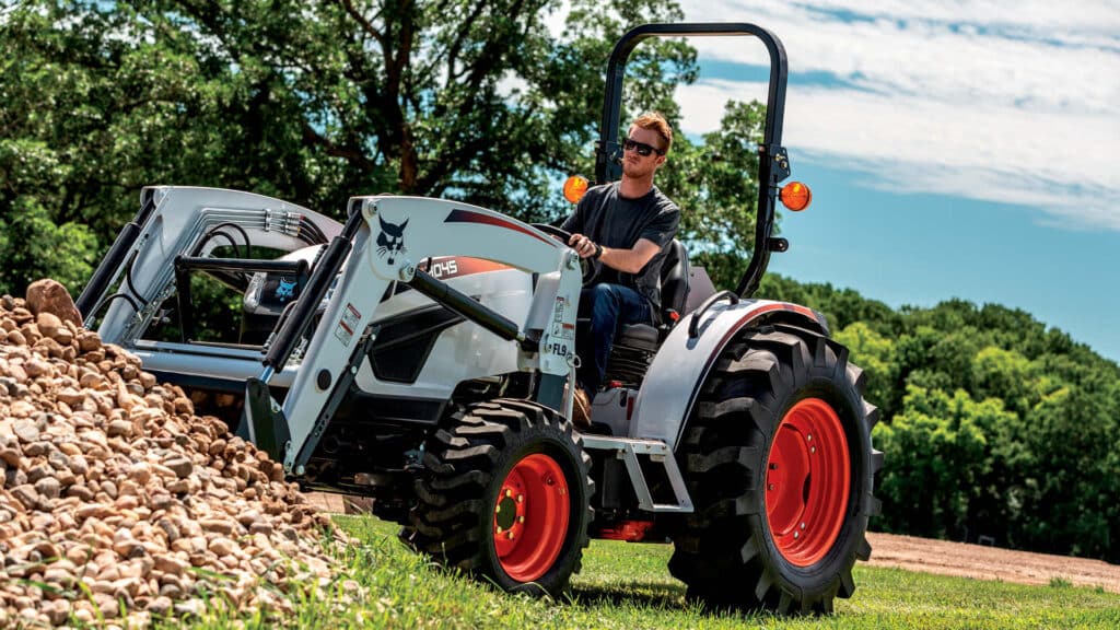 Man operating a small tractor for sale with front-end loader, moving a pile of rocks on a sunny rural property.