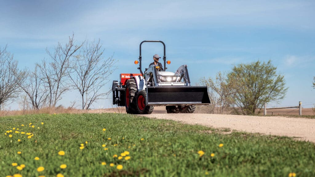 A man operates a Bobcat compact tractor with a front-end loader on a rural property, demonstrating how to choose the right compact tractor for tasks like moving soil and navigating uneven terrain.
