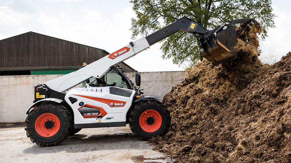 Bobcat TL38.70HF telehandler for sale moving a large pile of manure on a farm, highlighting power and hydraulic performance for heavy agricultural work.