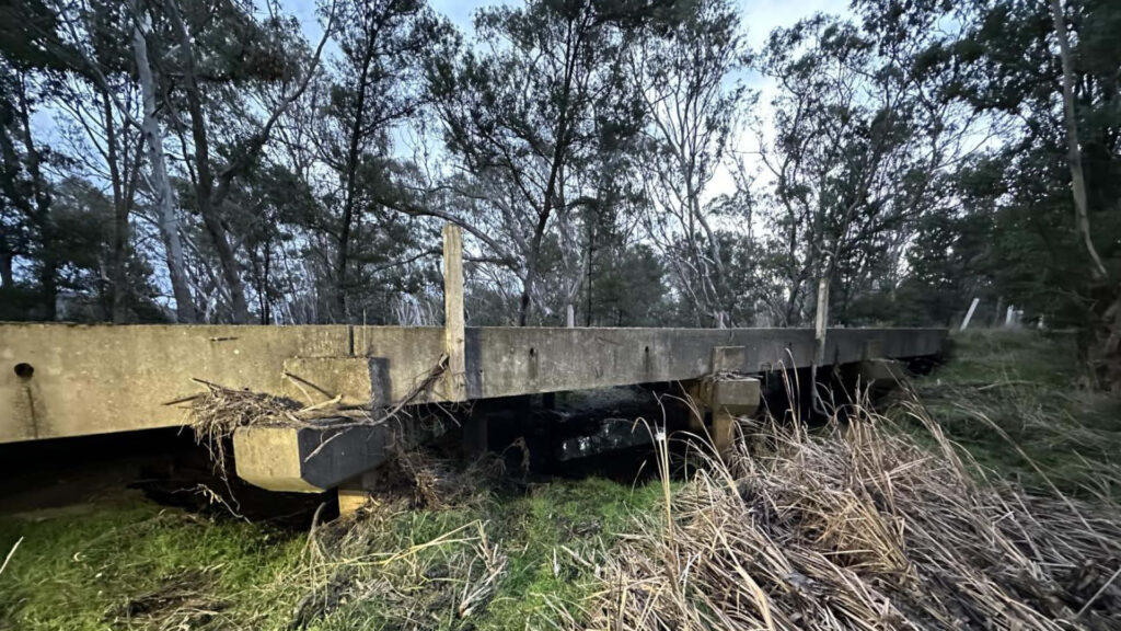 Low concrete Buangor–Ben Nevis road bridge with exposed piers, debris and eroded banks — the existing structure due for replacement.