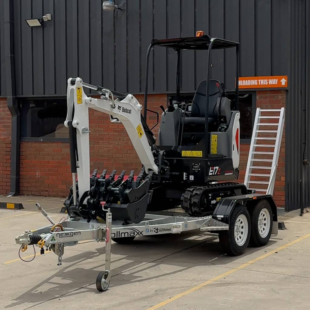 Bobcat E17 mini excavator trailer package loaded on a Rollmax trailer, showing the machine, bucket set, and ramps ready for transport at a worksite.