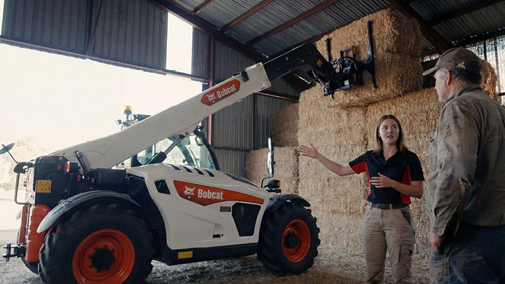 Delecca's sales specialist explaining Bobcat telehandler boom extension and lifting capability to farmer in agricultural shed with hay bales, demonstrating features that affect telehandler pricing and performance