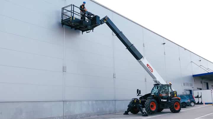 Bobcat construction telehandler with boom fully extended lifting worker in man platform to industrial warehouse wall showing safe height access and compliance