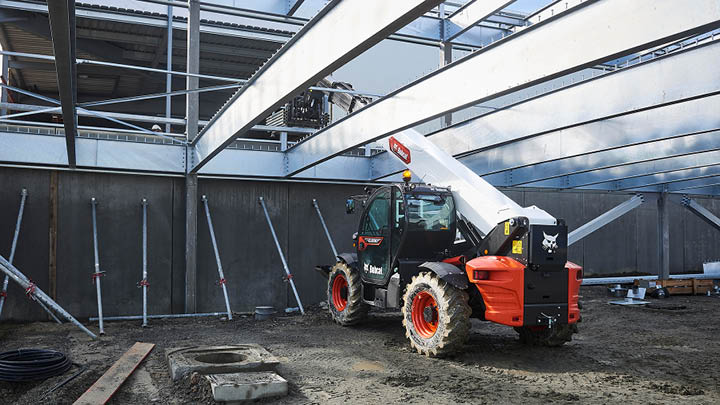 Bobcat T40.180 construction telehandler working inside a large steel frame industrial shed, boom extended to place materials at height