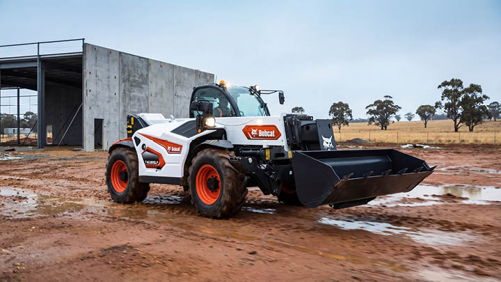 obcat T40.180SLP telehandler fitted with bucket attachment manoeuvring across a wet red clay construction site in Central Victoria, with a tilt-panel concrete building and eucalyptus trees in the background