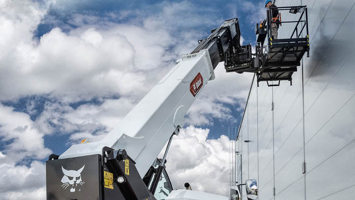 Bobcat T40.180SLP telehandler with man platform attachment elevated at height, with worker accessing a commercial building cladding facade against a dramatic cloudy sky