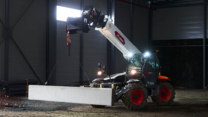 Bobcat telehandler with crane jib attachment lifting a concrete panel inside a large industrial shed at night, work lights illuminated
