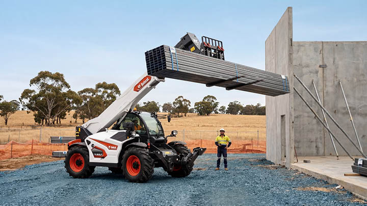 Bobcat T40.180 construction telehandler lifting a bundle of steel purlins at a tilt-panel concrete construction site in Central Victoria, with a Hi-Vis worker observing the lift