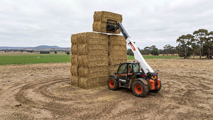 Bobcat TL43.80HF agricultural telehandler stacking large square hay bales to maximum height on an open Central Victorian farm, boom fully extended with Grampians ranges visible in background