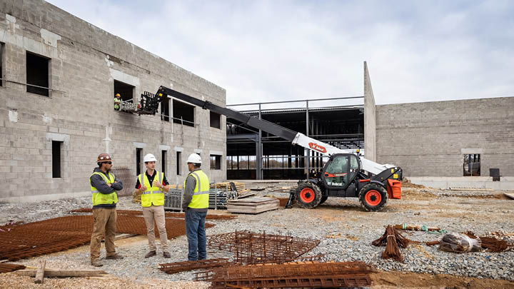Construction workers observing Bobcat telehandler on commercial building site with concrete structure and rebar showing lift height and reach capability