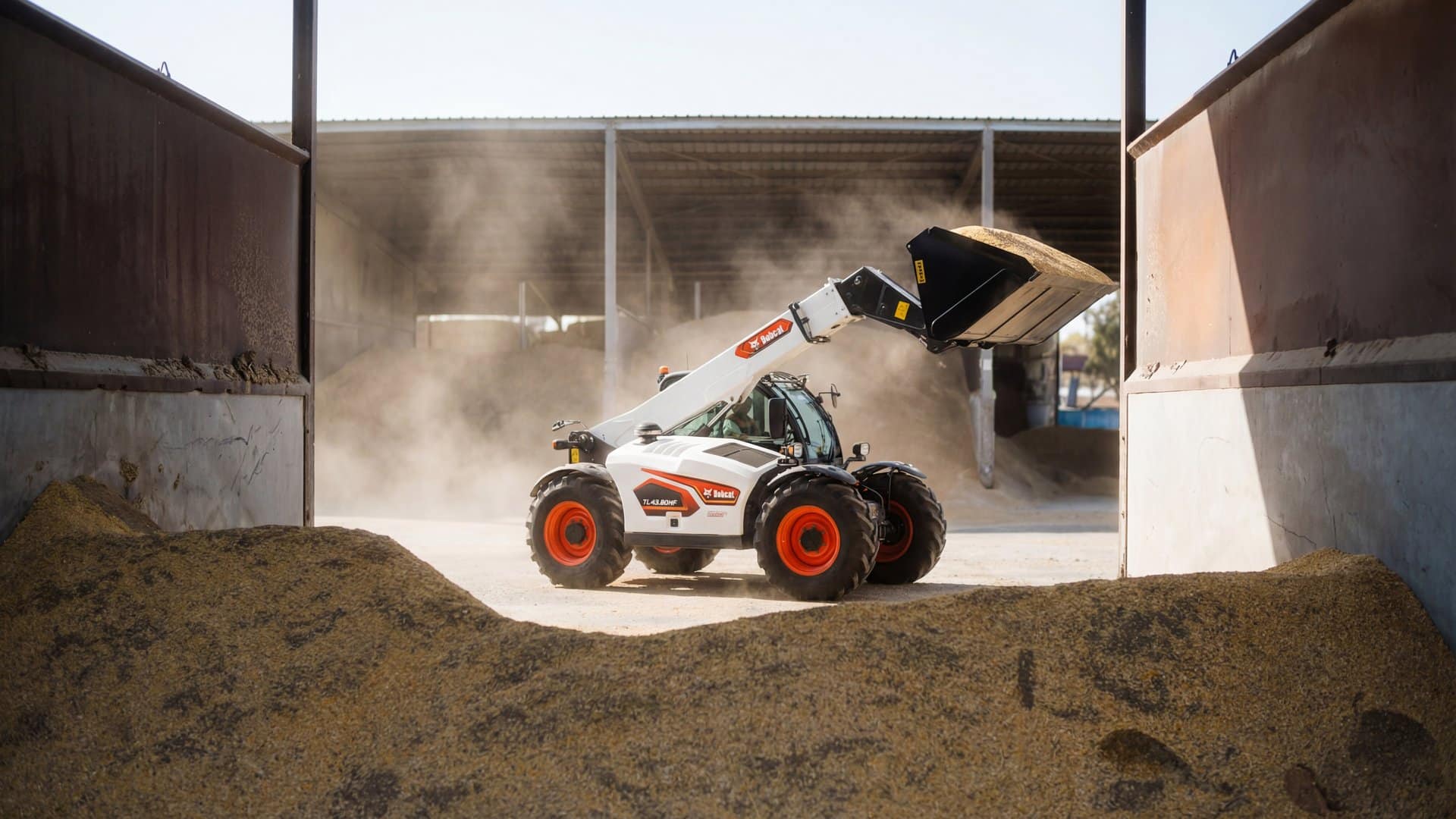 Bobcat TL43.80HF telehandler with commodity bucket raised, dumping grain inside a covered feedlot shed between concrete bunkers