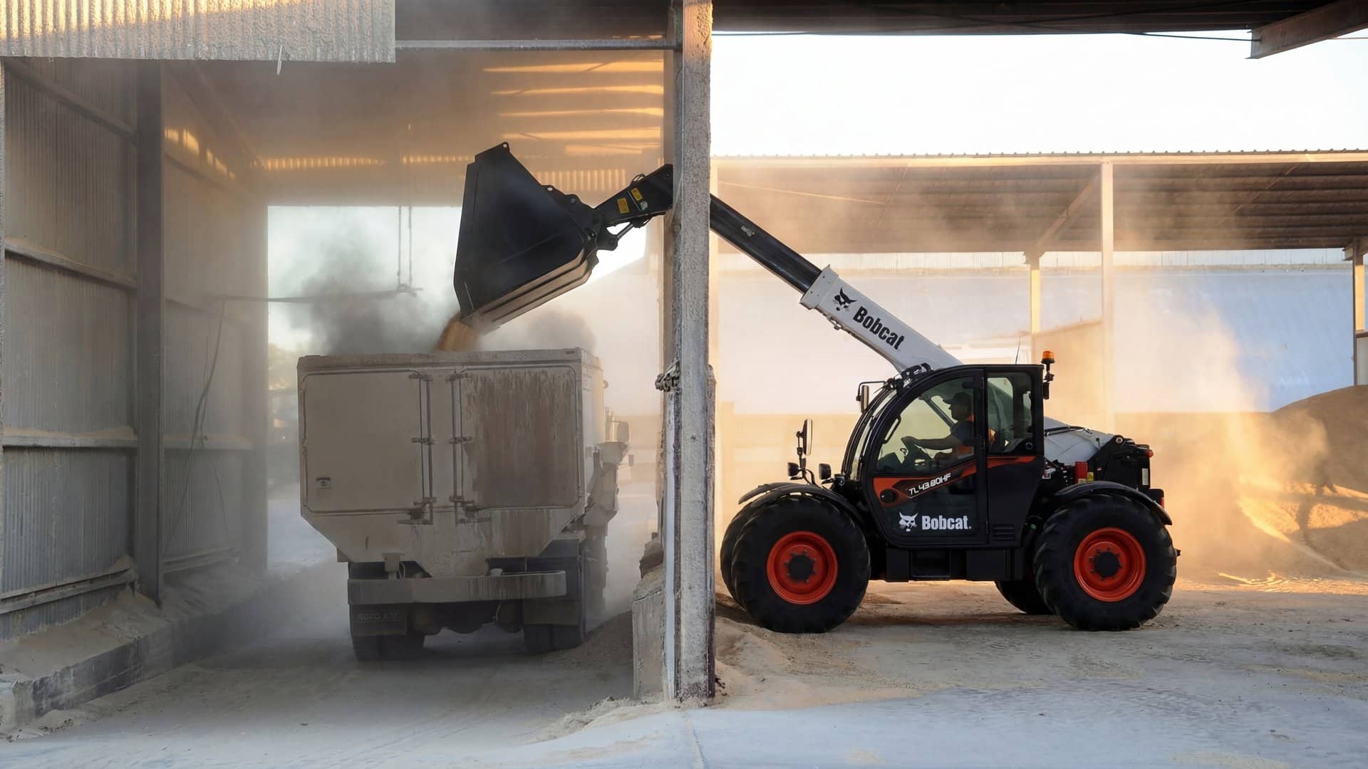 Bobcat TL43.80HF telehandler loading a feed mixer truck from a grain pile under a covered commodity shed
